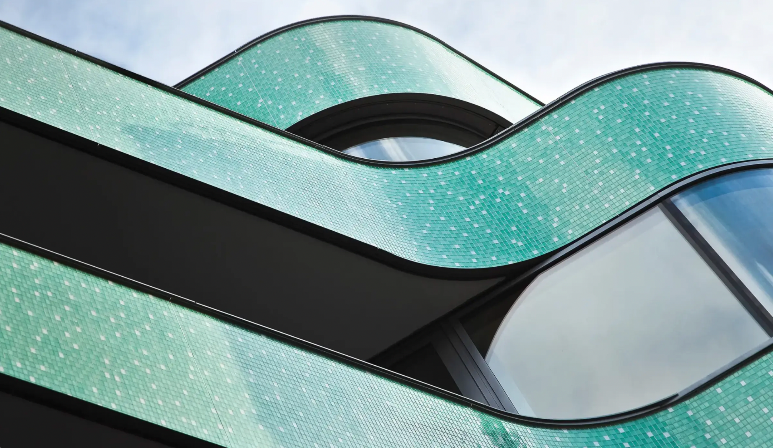 Wide-angle view looking up at swerving tiered balconies clad in aqua-colored Sto glass mosaic panels, illustrating dynamic architectural curves and high-performance rainscreen system designed for durability and aesthetic appeal in Atlantic Canadian climates.